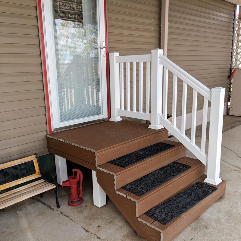 Elevate your manufactured home's entrance with a high-durability, low-maintenance entry solution. This small composite deck platform features premium brown decking with matching composite steps. A stark white low-maintenance vinyl railing system provides safe and stylish access, defined by clean vertical balusters and classic post caps. The set includes a robust handrail for the four-tread stair section.