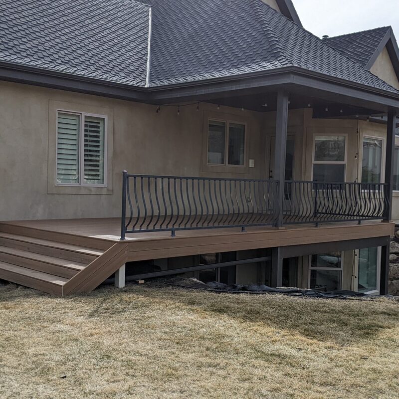 Expand your outdoor living possibilities with this custom deck by DeckPro LLC, featuring low-maintenance composite decking and a highly detailed railing system. The warm brown composite boards provide a durable, splinter-free surface, seamlessly transitioning to wide stairs that connect the home to the large yard. Sleek black metal railings with unique curved balusters wrap around the entire perimeter, offering a modern touch that contrasts beautifully with the home’s stucco exterior and brick accents. Pictured on a sunny day, this design illustrates how DeckPro can create a functional, stylish, and safe retreat for any home, regardless of terrain, thanks to a robust structural support system and expertly integrated rock retaining walls.