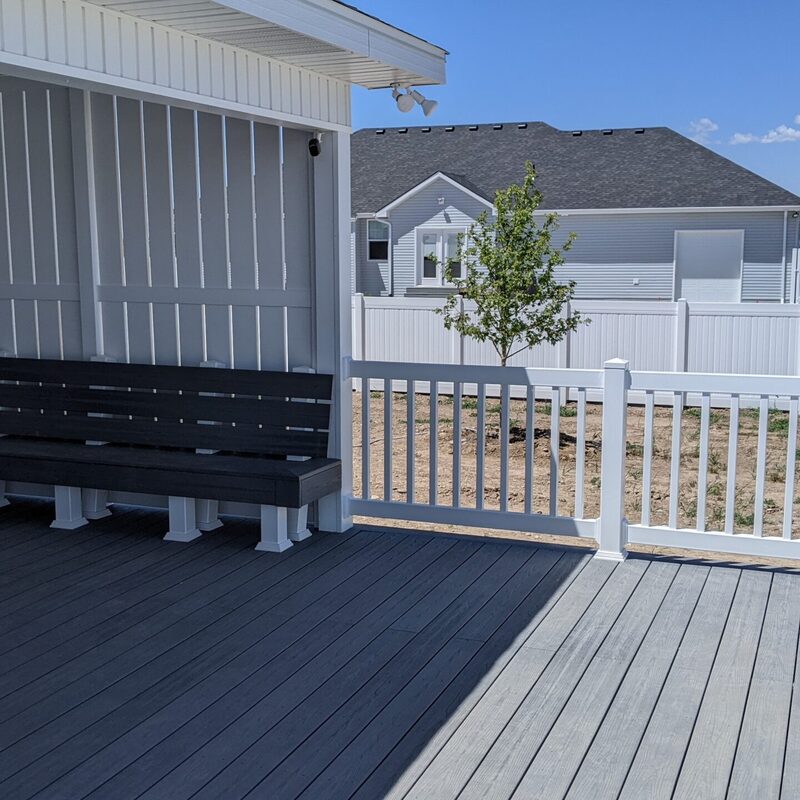 Expand your outdoor living possibilities with this extensive multi-level deck project by DeckPro LLC. Pictured on a sunny day, this build features high-quality composite decking in a warm wood-grain finish and contrasting dark perimeter trim. For the ultimate in style and function, we installed highly detailed black metal railings with unique curved balusters and wide, inviting stairs. The raised structure integrates seamlessly with natural rock retaining wall landscaping, leading down to a lower-level concrete patio for a truly multi-functional backyard retreat.