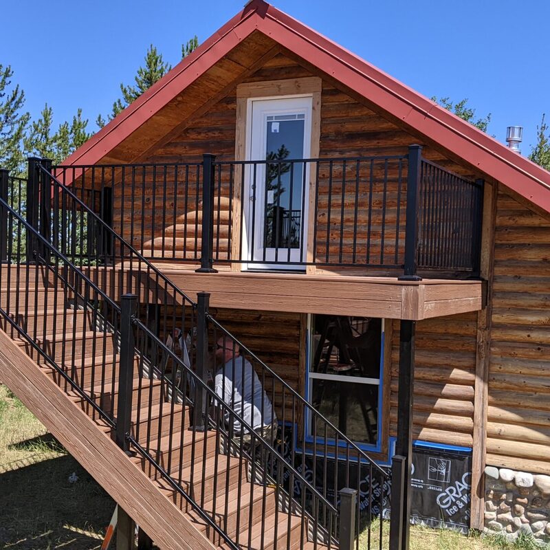 This aerial shot captures a custom project by DeckPro LLC designed to perfectly complement a rustic log cabin. The second-story composite deck features durable materials, sleek black metal railing, and a sturdy staircase connecting the upper level to the yard below.