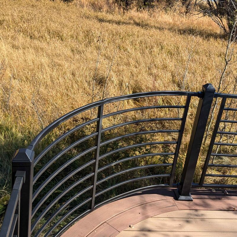 This elevated view showcases a custom curved Trex deck, expertly framed by a sleek and modern curved aluminum cable railing system. This hybrid design features black aluminum posts and rails with horizontal stainless steel cable infill, providing both safety and unobstructed views of the scenic, grassy mountain landscape in Sandy, Utah. The Trex composite decking in a warm brown tone (likely Transcend Havana or similar) perfectly complements the dark railing.