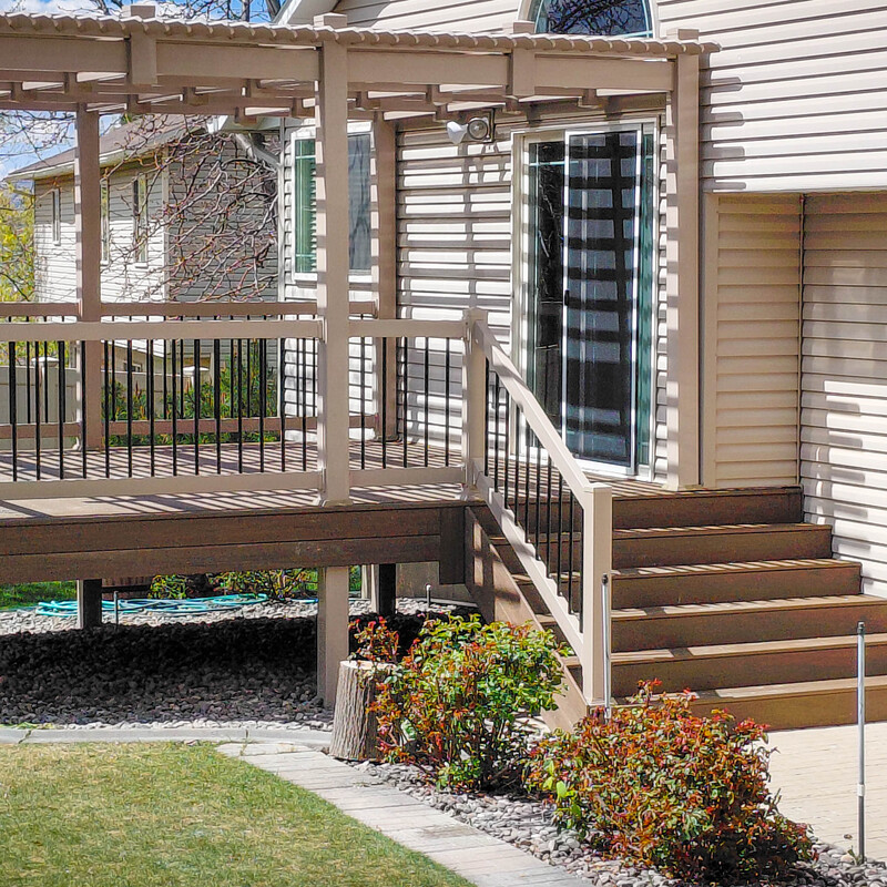 View from under a tan patio cover on a brown composite deck with black metal railings, featuring a sliding glass door and outdoor lighting.
