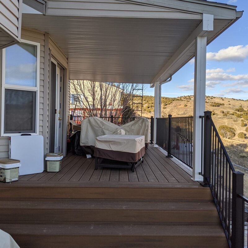 Take a step up into complete comfort with this covered composite deck by DeckPro LLC. This design seamlessly connects a lower patio to a covered upper-level living space via a wide staircase, providing an expansive area for relaxing and entertaining. The integrated roof cover offers reliable shelter from the elements, while sleek black metal railings define the perimeter, blending modern safety with a rustic mountain-side view.
