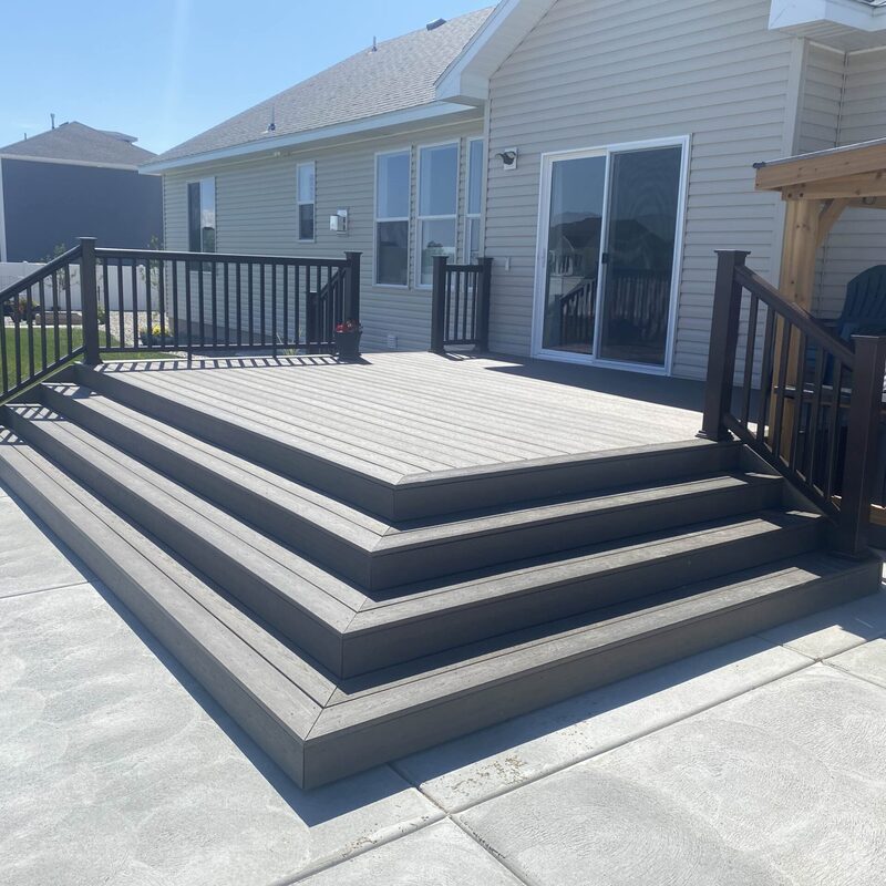 Expand your outdoor living possibilities with this extensive composite deck by DeckPro LLC, beautifully attached to a classic log home cabin. This custom raised structure transitions seamlessly into the natural, tree-filled landscape, providing the perfect viewpoint. We installed high-quality, low-maintenance composite decking in a warm brown tone and paired it with sophisticated black metal perimeter railings that complement the rustic aesthetic. Pictured on a beautiful day, the build showcases distinct railing shadows, creating a sturdy and stylish retreat perfect for relaxation or entertaining.