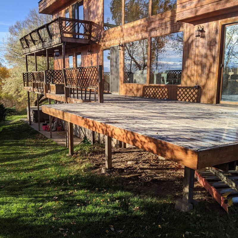 Extend your upper-level living space with this cozy raised fir balcony deck by DeckPro LLC, which combines rustic elements with modern comfort. We installed extensive natural wood decking and a complete perimeter railing system featuring a continuous top rail and balusters, attached to a house with clean white horizontal siding. Pictured on a sunny day with distinct shadows cast across the deck boards, this substantial build offers tranquility and function, overlooking a natural landscape with a dirt road, pine trees, and a basketball hoop—the perfect mountain retreat to take in scenic woodland views.