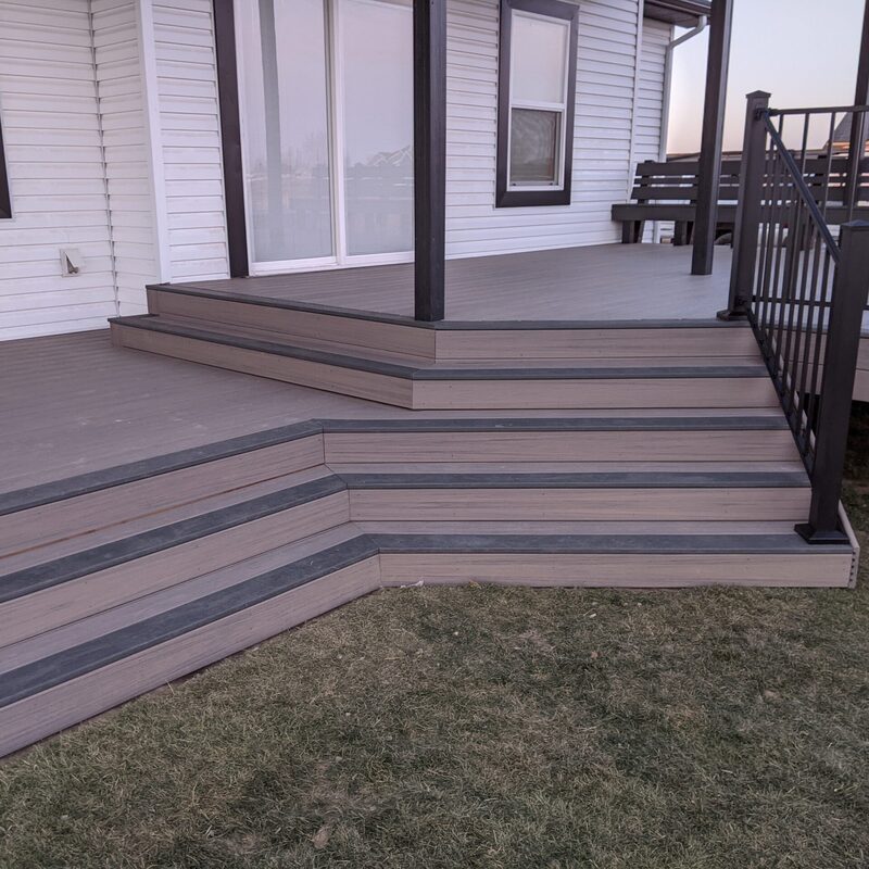 Make a grand entrance to your backyard with custom craftsmanship from DeckPro LLC. This grey composite deck features elegant, wide cascading stairs highlighted by dark "picture framing" borders and sleek black metal railing, perfect for Southeast Idaho homes.