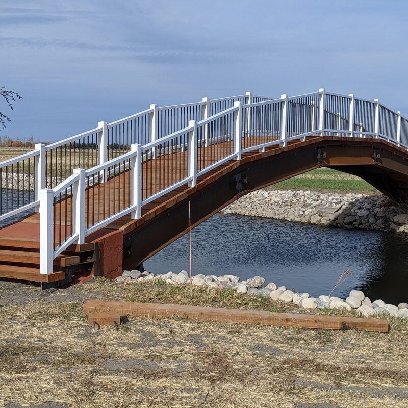 Experience unparalleled access across challenging terrain with this custom-engineered pedestrian bridge by DeckPro LLC. This elevated footbridge is a masterpiece of design and durability, designed to span natural and man-made water features. Crafted with premium multi-tonal grey grooved composite decking boards for a long-lasting and anti-slip walking surface, it is safely enclosed by a safe, multi-material railing system.