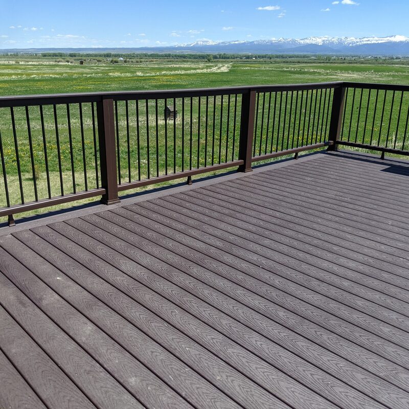 Discover The Range View Composite Deck with Panoramic Mountain Vista, a signature elevated custom deck built by the experts at DeckPro LLC. This expansive project features high-quality, grooved, low-maintenance composite decking boards in a multi-tonal grey and brown mix, providing a durable and stylish walking surface. Securely enclosed by a matching dark composite and metal baluster railing system, the deck is custom-designed to perfectly frame the stunning, uninterrupted panoramic views over a lush green meadow, rolling hills, and the distant, snow-capped mountain range under a clear blue sky.
