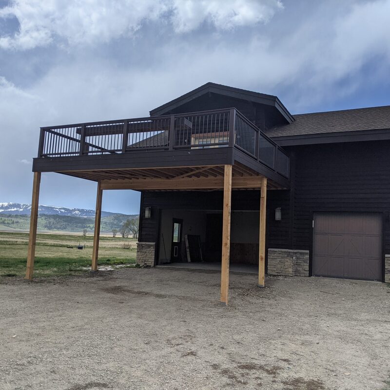A wide-angle exterior photograph from a gravel driveway showing a large, custom-engineered elevated multi-bay garage with two bays, integrated into a dark brown board and batten siding lodge-style cabin on a hillside. A massive, second-story covered living deck, built by DeckPro LLC, spans the length of the garage.