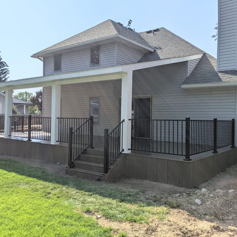 Extend your multi-level outdoor living with this dramatic multi-zone composite raised deck transformation by DeckPro LLC. Pictured on a beautiful partly cloudy day, this sophisticated structure features expansive gray and brown composite planks paired with sleek black vertical perimeter railings. This sturdy retreat transitions via extensive massive wide staircases with contrasting risers to a stamped concrete lower level and a beautifully landscaped rock garden featuring natural boulder rocks on a gravel bed. Direct access point via multiple existing glass doors and a large massive set of connecting stairs allow for effortless entertaining, while integrated recessed ceiling lighting in the covered upper area ensures evening use. Defining functional zones perfect for both relaxation and large-scale entertaining, this unique tranquilidad sanctuary maximizes spectacular multi-directional natural views.