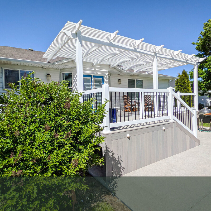 Captured from the yard, this image highlights the structural integration of a custom DeckPro LLC build. The elevated light grey composite deck flows perfectly out of the home's back doors, sheltered by a robust white pergola that is securely attached to the roof eave. The deck's understructure is cleanly hidden using matching vertical composite fascia boards, creating a polished, high-end look. A wide staircase provides easy, secure access down to the existing concrete patio.
