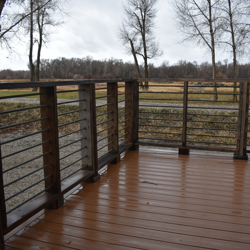 An on-deck view of a custom rustic outdoor living space built by DeckPro LLC. This deck features warm brown composite decking engineered to withstand wet weather and natural elements without rotting. The unique, custom-built railing system utilizes sturdy dark wood posts paired with horizontal metal rods, creating a rugged, industrial-cabin aesthetic that perfectly complements the surrounding natural landscape.