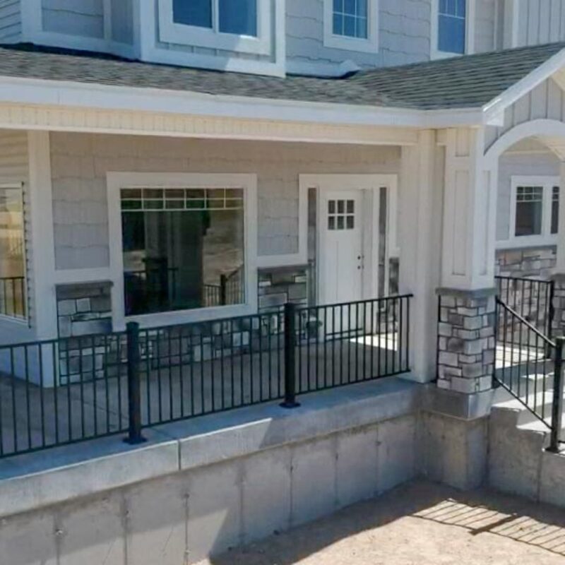 Welcome to our signature The Crown Jewel, an elevated custom-built front porch by DeckPro LLC, transforming the entrance of a modern custom home on difficult, sloped topography. This monumental covered concrete platform completely redefines the home's primary entry. Crafted with durable black metal picket railings (balustrades) with vertical pickets, matching posts, and continuous handrails, this raised porch offers low-maintenance, sophisticated safe-access.