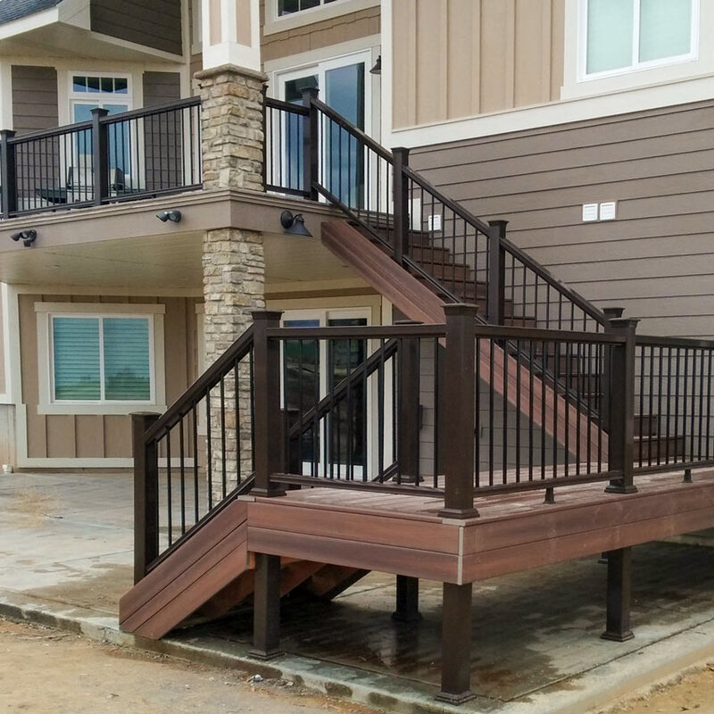 Elevate your home's outdoor living space with this custom multi-level deck project by DeckPro LLC. Featuring durable composite decking in a warm tone, the design connects an upper-level balcony to a lower-level deck and patio via a grand staircase. Dark metal railings and beautiful stone veneer columns add a touch of elegance and structural integrity to this impressive backyard upgrade.