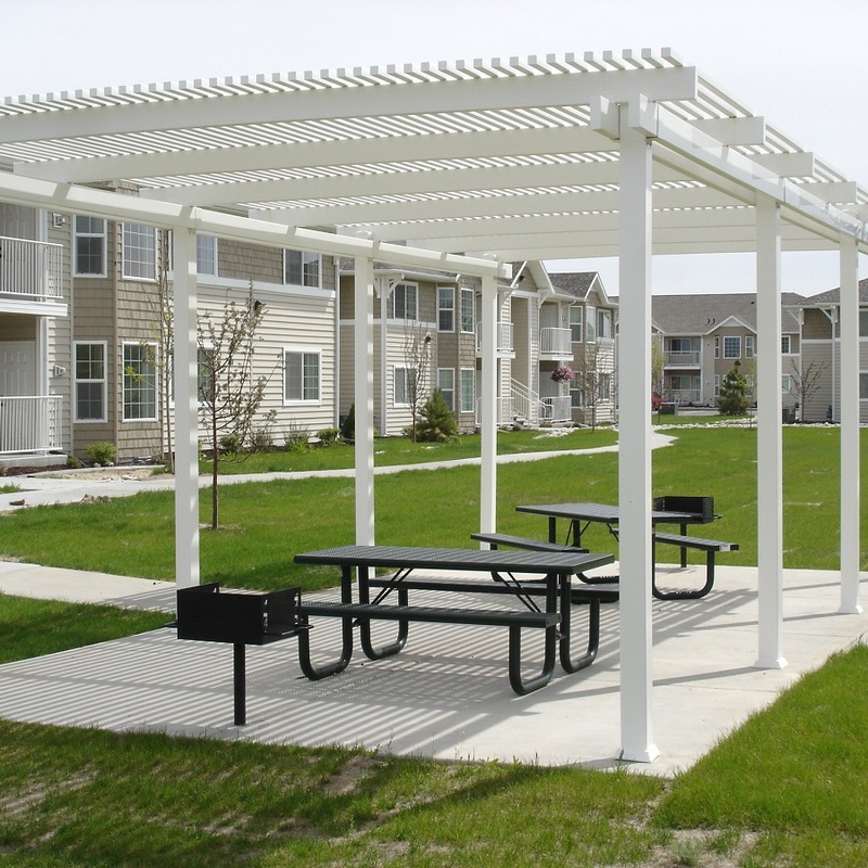 A photograph of a large, custom-engineered, multi-bay white vinyl louvered pergola pavilion installed in the common area courtyard of a multi-story apartment complex. This free-standing structure, crafted by DeckPro LLC, covers a concrete pad that features two commercial-grade perforated metal picnic tables and two permanent charcoal grills.